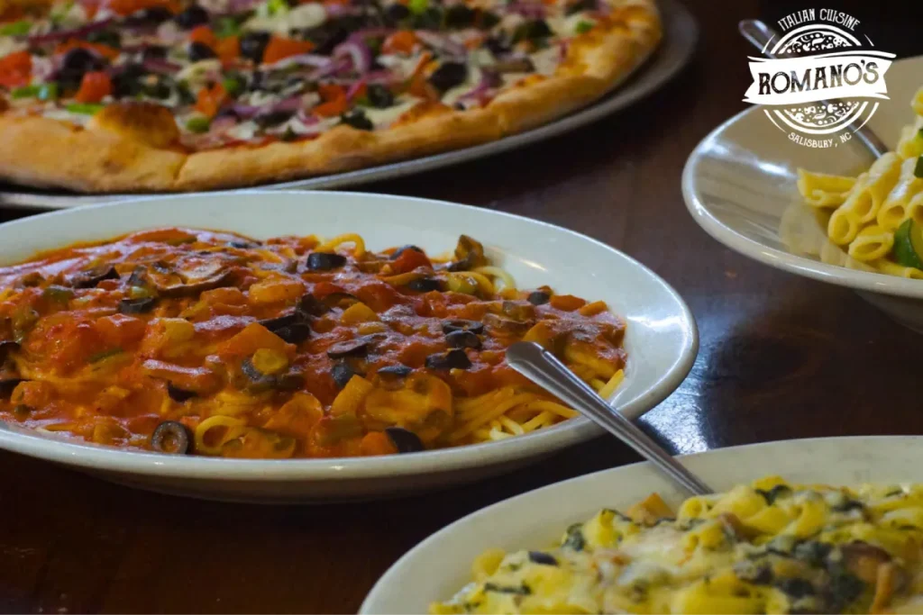 A plate of pasta with tomato sauce, olives, and vegetables, with pizza in the background.